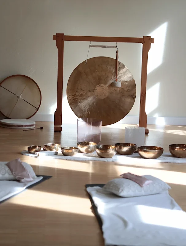 Ein sonnendurchfluteter Raum, vorbereitet für eine Klangmeditation. Im Zentrum steht ein großer Gong in einem Holzrahmen, davor sind mehrere goldene Klangschalen auf dem Boden aufgereiht. Im Vordergrund liegen zwei Yogamatten mit Kissen und Decken bereit.