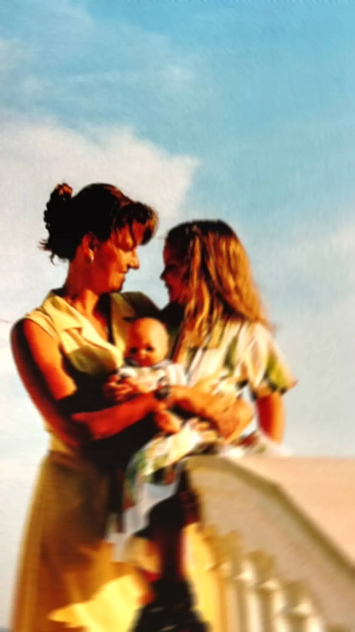 Eine Mutter hat ihren Arm um ihre Tochter gelegt, die sitzend auf einer Brüstung eine Baby-Puppe hält. Die Personen befinden sich im Freien vor einem blauen Himmel im warmen Sonnenlicht.