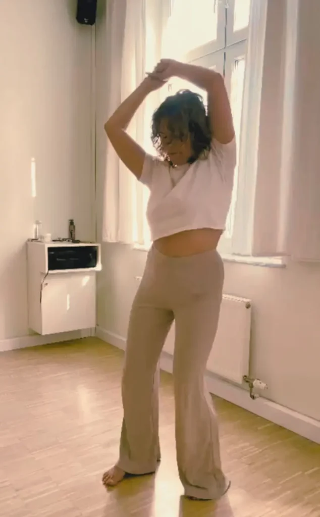 Eine Aufnahme in voller Länge einer Frau, die barfuss tanzt, ihre Arme über ihrem Kopf, in einem lichtdurchfluteten Innenraum mit Holzboden und einem Fenster im Hintergrund.