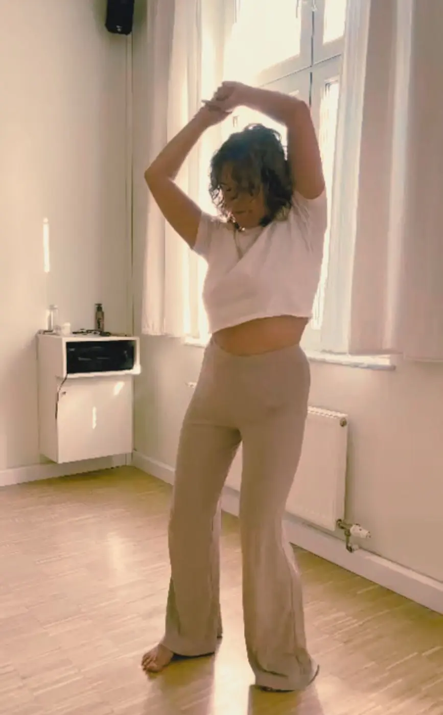 Eine Aufnahme in voller Länge einer Frau, die barfuss tanzt, ihre Arme über ihrem Kopf, in einem lichtdurchfluteten Innenraum mit Holzboden und einem Fenster im Hintergrund.