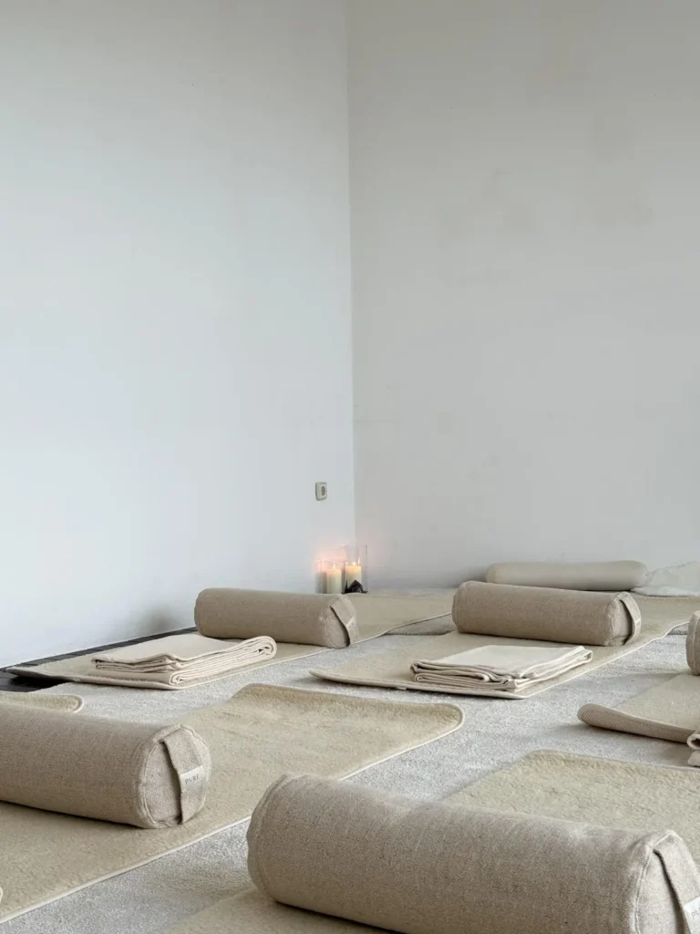 Ein heller, minimalistischer Raum, der für Yoga oder Meditation vorbereitet ist. Auf dem Boden liegen mehrere cremefarbene Matten, die jeweils mit einer zylindrischen Nackenrolle und einer gefalteten Decke ausgestattet sind. In der Ecke brennen gemütlich Kerzen.