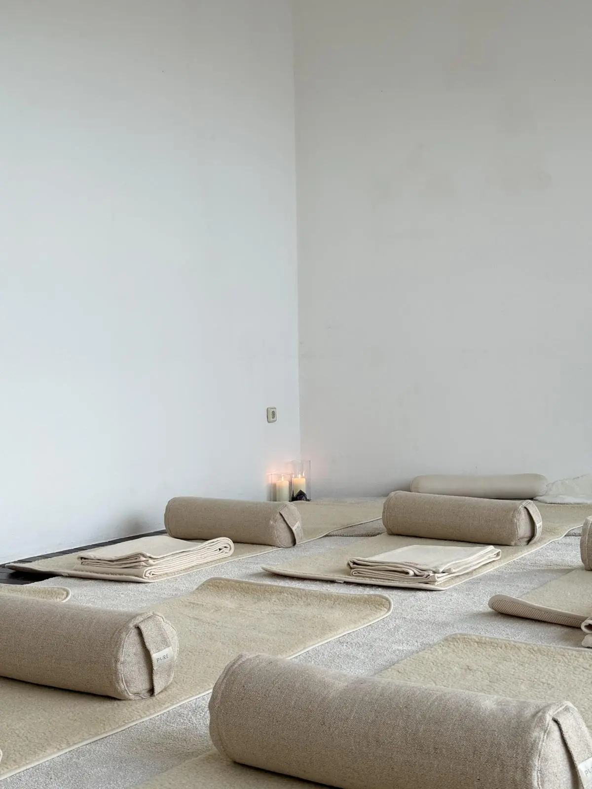 Ein heller, minimalistischer Raum, der für Yoga oder Meditation vorbereitet ist. Auf dem Boden liegen mehrere cremefarbene Matten, die jeweils mit einer zylindrischen Nackenrolle und einer gefalteten Decke ausgestattet sind. In der Ecke brennen gemütlich Kerzen.