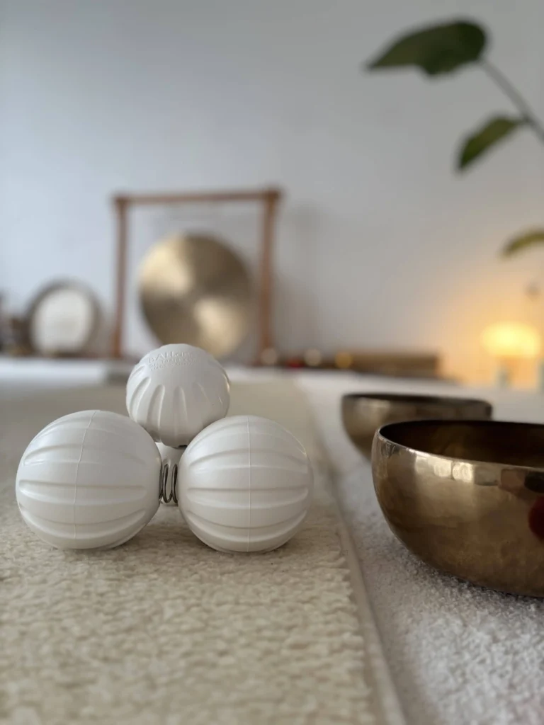 Eine Nahaufnahme zeigt drei weiße, strukturierte Faszien-Massagebälle in einer dreieckigen Formation auf einer flauschigen, cremefarbenen Teppichmatte. Rechts daneben stehen zwei polierte Messing-Klangschalen.