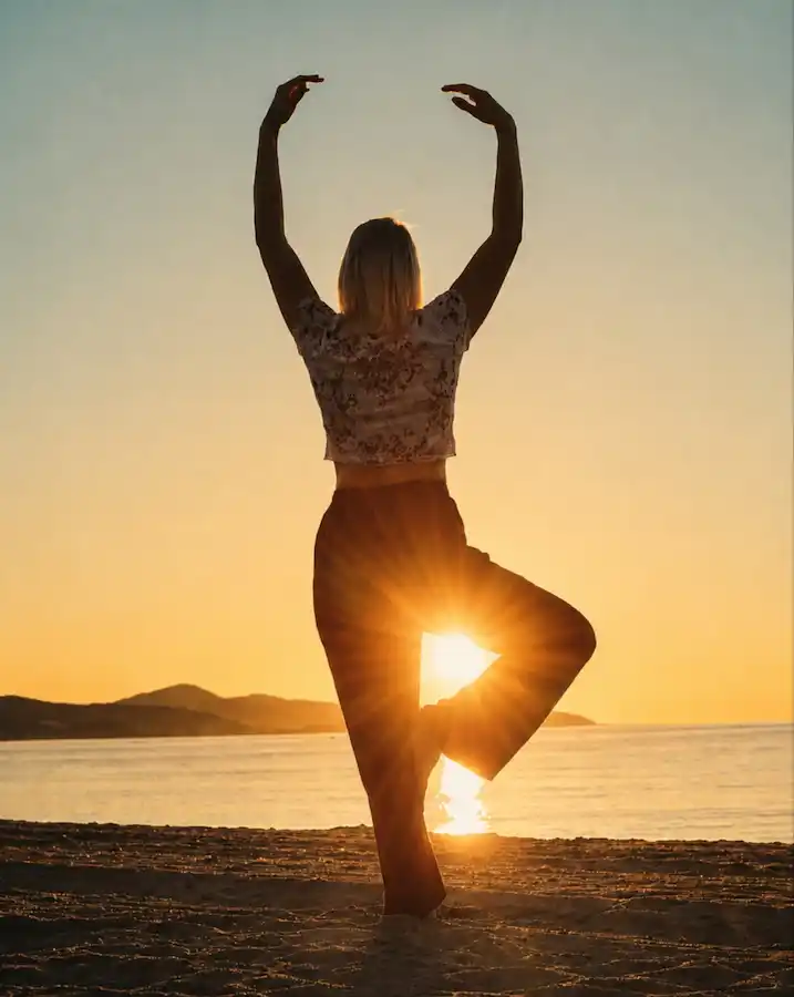 Eine Frau von hinten fotografiert in einer Yoga-Haltung am Strand bei Sonnenuntergang. Die tiefstehende Sonne scheint genau zwischen ihren Beinen hindurch.