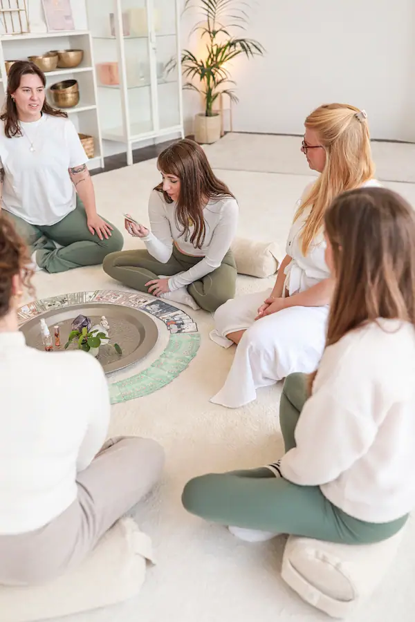 Gruppe von Frauen sitzt im Kreis auf Kissen, in der Mitte eine runde Schale und drumherum Karten ausgelegt. Eine Teilnehmerin hält eine Karte in der Hand und liest den anderen daraus vor.