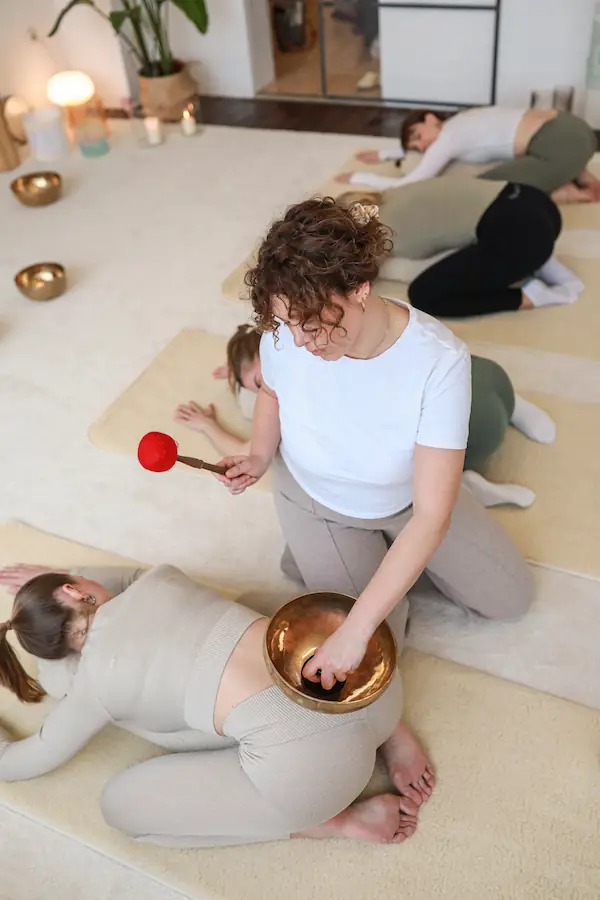 Elina Lumida spielt mit einem Klangschlägel auf einer Klangschale, die am unteren Rücken einer Kursteilnehmerin aufgestellt ist. Weitere Teilnehmerinnen liegen im Hintergrund auf Matten in der Kindspose.