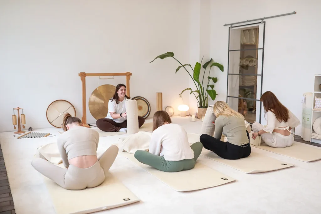 Lachende Frau sitzt vor einem Gong und hält einen vor sich aufgestellten Yogakissen, während Teilnehmerinnen, die auf Matten ihr gegenüber sitzen, ihr nachahmen.