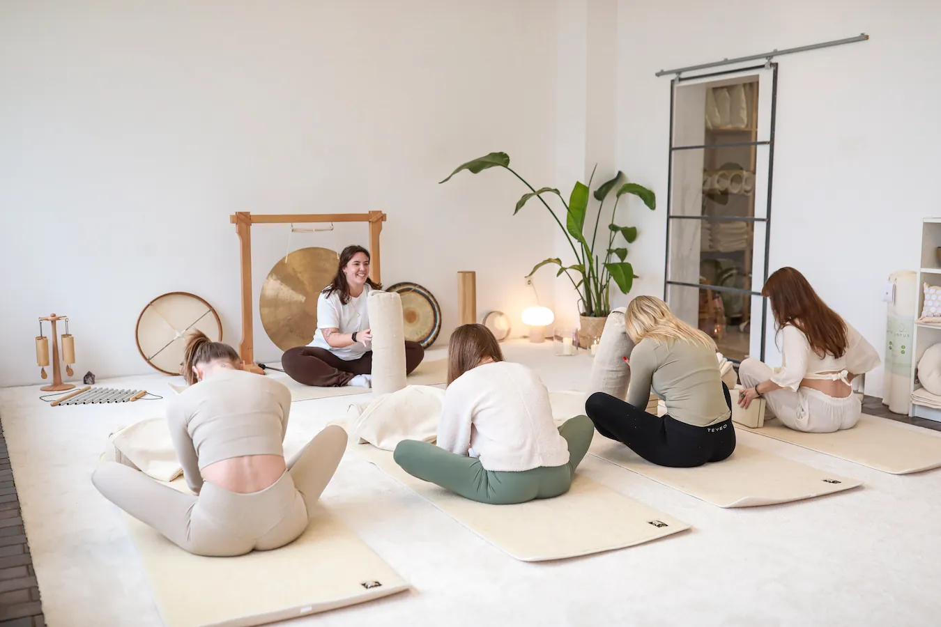 Lachende Frau sitzt vor einem Gong und hält einen vor sich aufgestellten Yogakissen, während Teilnehmerinnen, die auf Matten ihr gegenüber sitzen, ihr nachahmen.