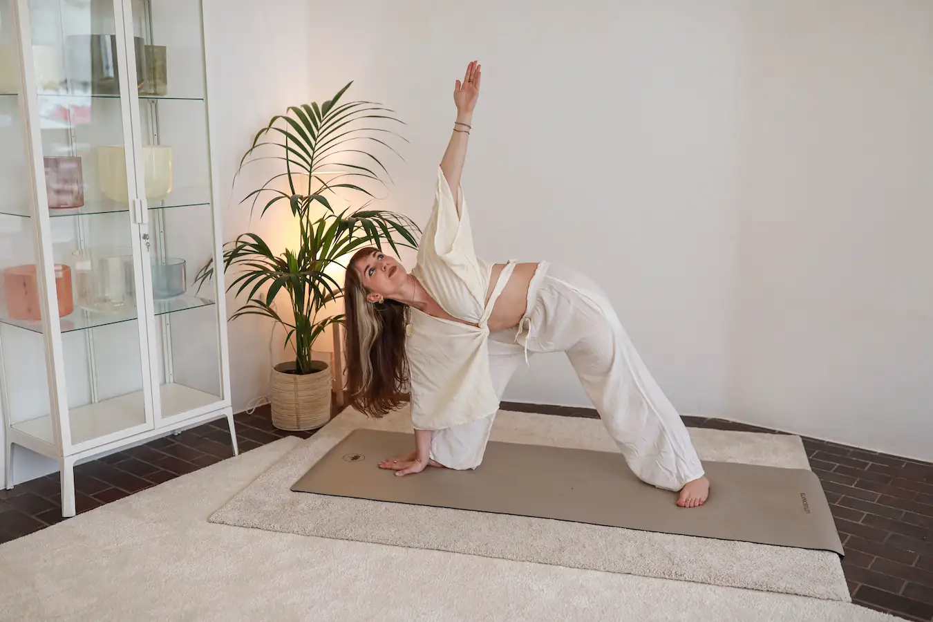 Frau in cremefarbener Kleidung übt eine Yoga-Dehnübung mit ausgebreiteten Beinen und ausgestrecktem Arm auf einer Matte